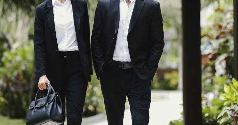 Two people in black suits walk side by side outdoors; one carries a black leather briefcase.