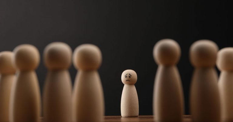 A small wooden peg figure with a sad face stands alone among taller figures in a dark setting.