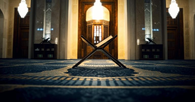 Open Quran stand (rehal) with an open book in a mosque, centered on a patterned prayer rug with warm hanging lights overhead