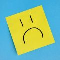 Yellow sticky note with a simple sad face drawn in black on a blue background.