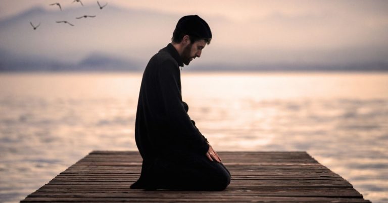 Man kneeling in prayer on a wooden pier at sunset over calm water.