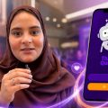 A woman in a brown hijab demonstrates the Think Quran app on a large phone screen with the app's robot logo and purple background.