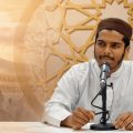 Man in a white robe speaks into a microphone at a desk, with a gold geometric backdrop and a Kaaba scene in the background.