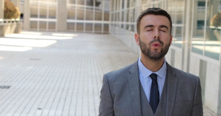 Man in a gray suit outdoors, eyes closed, lips puckered as if blowing a kiss toward the camera.