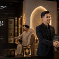 Two men in business attire shake hands in a showroom with Islamic decor and a sign that reads 'Peluang Ejen' in the background.