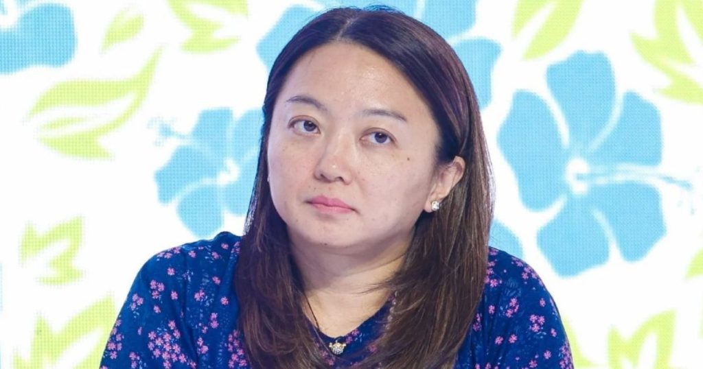 Woman with long brown hair wearing a blue floral blouse, looking slightly to the left against a bright floral backdrop.