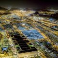 Aerial night view of a sprawling desert city with brightly lit grid-like housing blocks and winding roads.