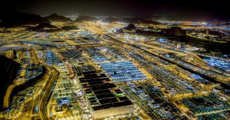 Aerial night view of a sprawling desert city with brightly lit grid-like housing blocks and winding roads.