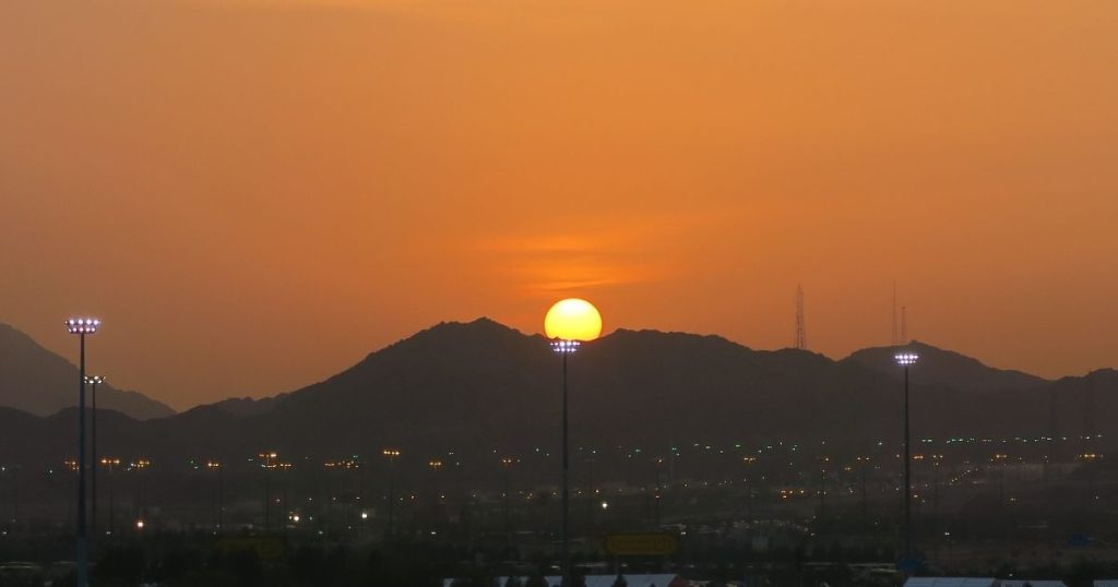 Sun setting behind a silhouette of mountains with tall stadium lights in the foreground and city lights below, creating a warm orange glow.