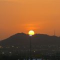 Sun setting behind a silhouette of mountains with tall stadium lights in the foreground and city lights below, creating a warm orange glow.