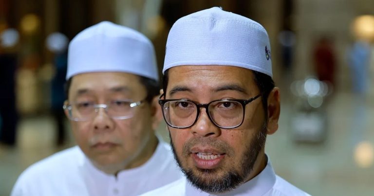 Close-up of a man with glasses and beard wearing a white cap, speaking, while another man in a white cap looks on in a clinical setting.
