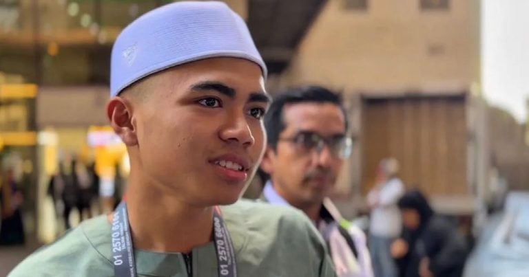Young man in light green medical scrubs and blue cap, outdoors with a blurred crowd behind him.