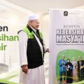 Two men in white traditional dress and green vests stand beside banners promoting a Hajj cleanliness campaign, with mosque silhouettes in the background.