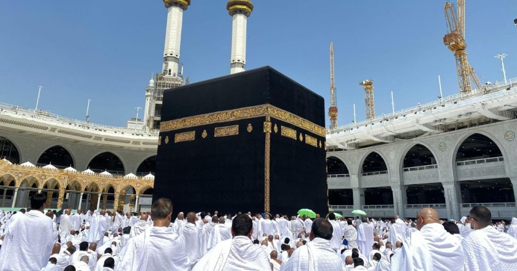 Pilgrims in white ihram surround the black Kaaba at the Grand Mosque in Mecca during pilgrimage under a blue sky.