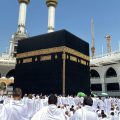 Pilgrims in white ihram surround the black Kaaba at the Grand Mosque in Mecca during pilgrimage under a blue sky.