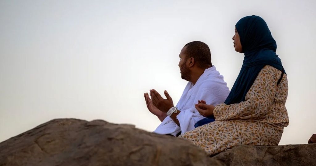 Man and woman in traditional clothing sit on rocks, praying with raised hands outdoors.