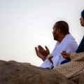 Man and woman in traditional clothing sit on rocks, praying with raised hands outdoors.