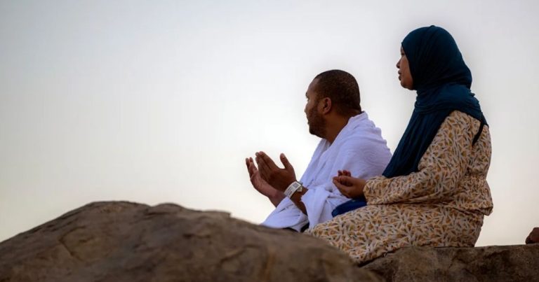 Man and woman in traditional clothing sit on rocks, praying with raised hands outdoors.