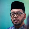 Man wearing a black traditional cap and glasses, with a short beard, speaking in a blue-green background.