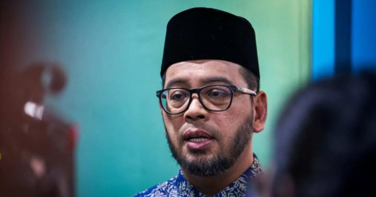 Man wearing a black traditional cap and glasses, with a short beard, speaking in a blue-green background.