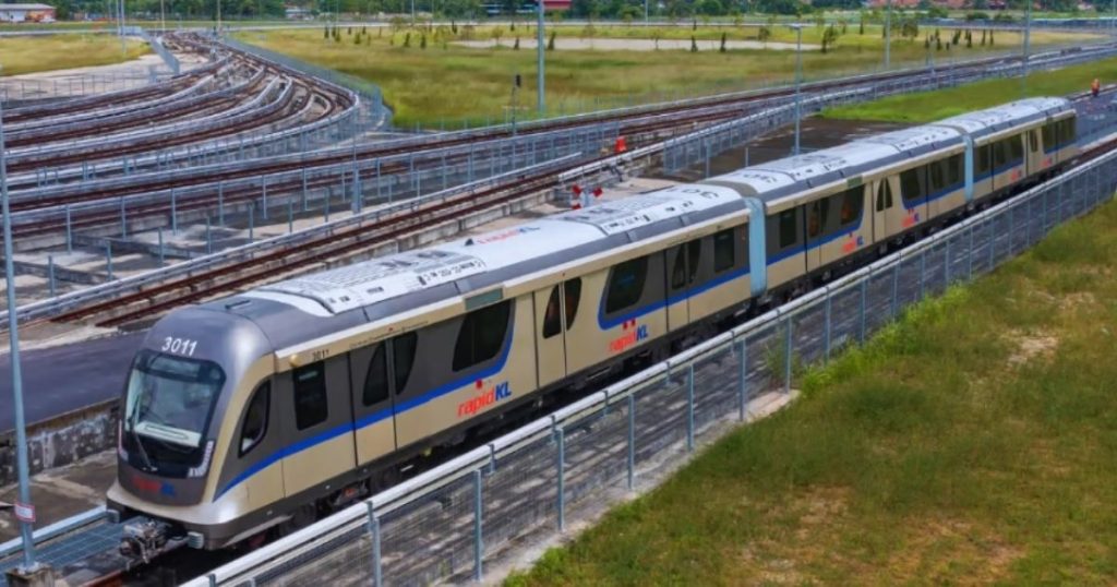 Modern silver-beige rapidKL passenger train on elevated tracks, curving rail lines in the background and green fields around the station area.