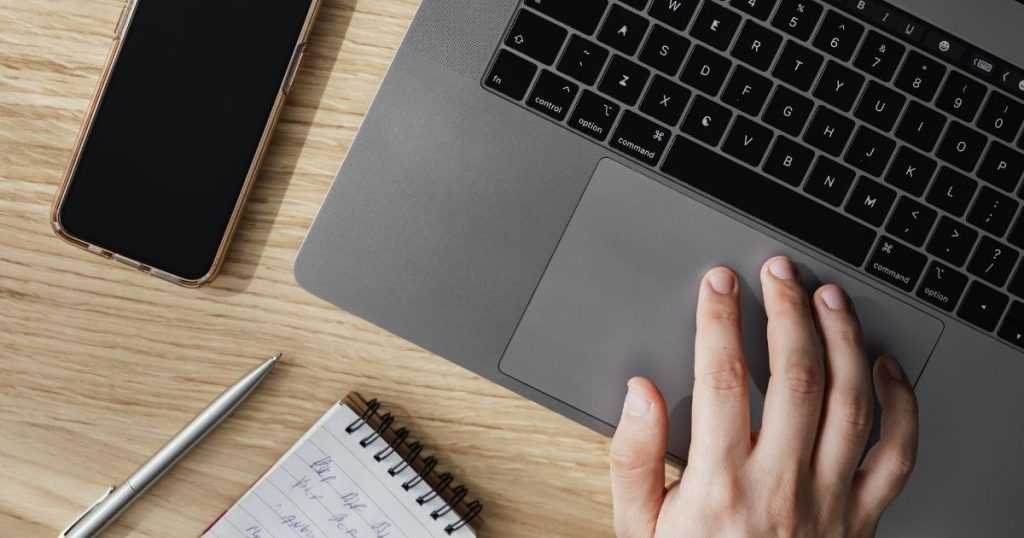 Person uses a laptop, resting a hand on the trackpad beside a notebook and phone on a wooden desk