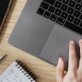 Person uses a laptop, resting a hand on the trackpad beside a notebook and phone on a wooden desk