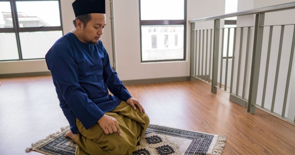 Man wearing a blue traditional outfit and prayer cap kneels on a patterned prayer rug, performing Muslim prayer indoors near a railing and windows.