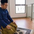 Man wearing a blue traditional outfit and prayer cap kneels on a patterned prayer rug, performing Muslim prayer indoors near a railing and windows.