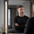 Man in a black shirt stands with arms crossed, looking at his reflection in a tall, framed mirror in a modern, softly lit room.