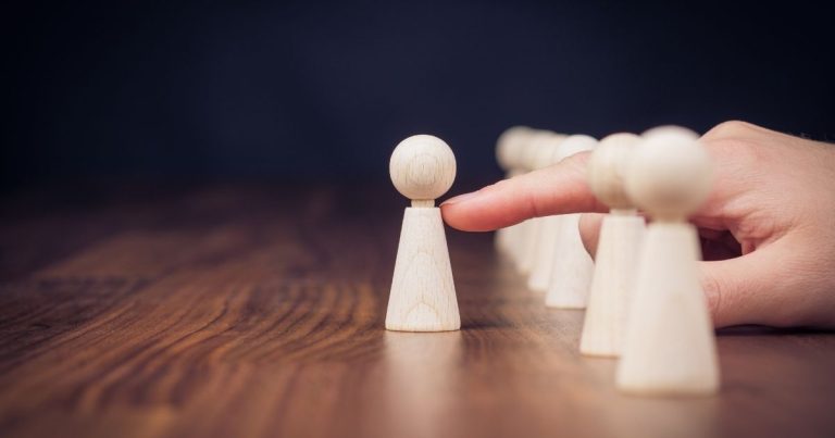 A wooden pawn at the front is touched by a finger, with a line of pawns behind it on a wooden table.