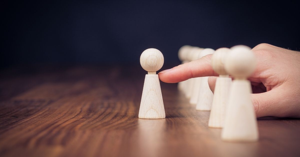 A wooden pawn at the front is touched by a finger, with a line of pawns behind it on a wooden table.