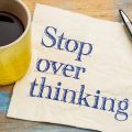Yellow mug of coffee beside a napkin with the phrase 'Stop over thinking!' and a black pen on a weathered blue wooden table.