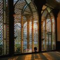 Sunlight filters through colorful stained-glass arches in a decorative hall, casting long warm shadows on the patterned floor.