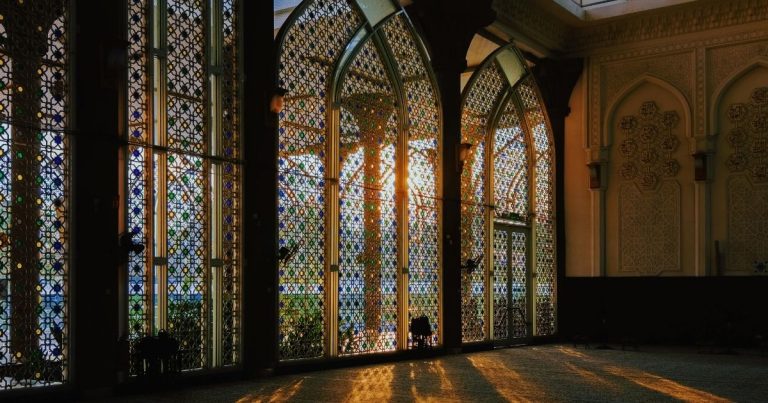 Sunlight filters through colorful stained-glass arches in a decorative hall, casting long warm shadows on the patterned floor.