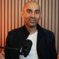 Bald man speaking into a microphone in a studio with orange lighting and vertical panel backdrop on the wall.