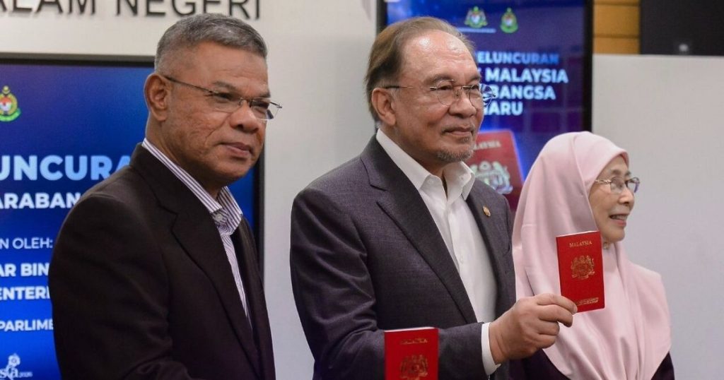 Three officials in suits and a pink-hijab-wearing woman hold red Malaysian passports at a formal launch event, with a banner in the background.