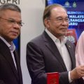 Three officials in suits and a pink-hijab-wearing woman hold red Malaysian passports at a formal launch event, with a banner in the background.