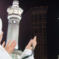 Muslim woman in a white hijab prays with hands raised, standing before illuminated twin minarets at night.