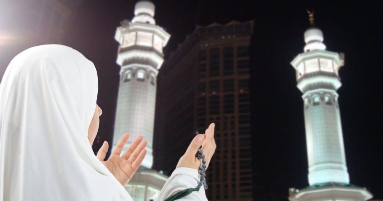 Muslim woman in a white hijab prays with hands raised, standing before illuminated twin minarets at night.