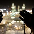 Silhouetted hands in prayer holding a string of beads against a blurred nighttime cityscape.