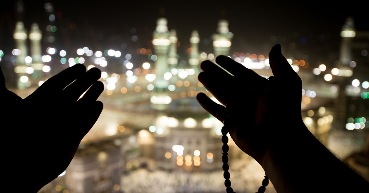Silhouetted hands in prayer holding a string of beads against a blurred nighttime cityscape.