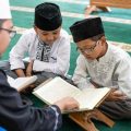 Imam guiding two boys as they read from the Quran on a green prayer rug in a mosque