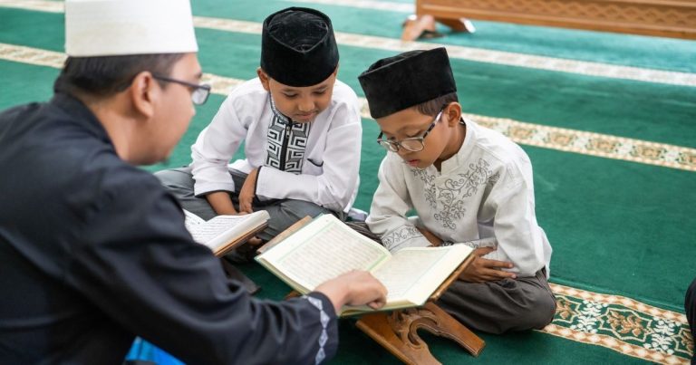 Imam guiding two boys as they read from the Quran on a green prayer rug in a mosque