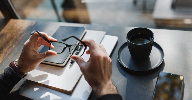 Hands hold eyeglasses over a notebook with a pen on a desk, beside a black coffee cup and a smartphone.