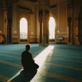 Silhouette of a person praying on a blue carpet inside a mosque, sunlight streaming through arched windows.