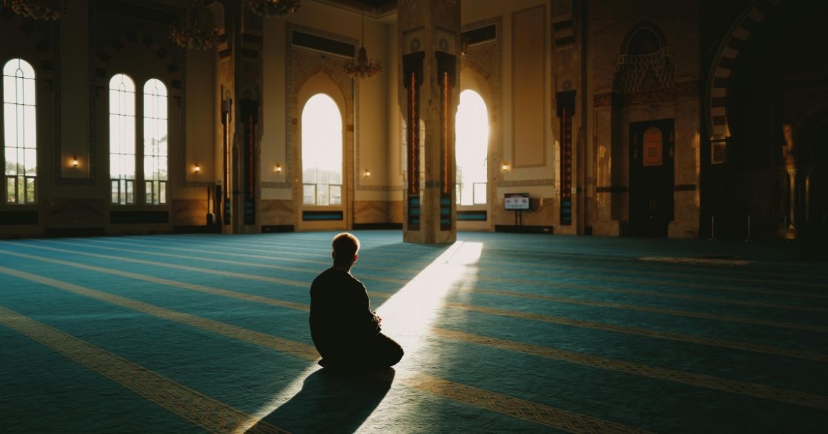 Silhouette of a person praying on a blue carpet inside a mosque, sunlight streaming through arched windows.