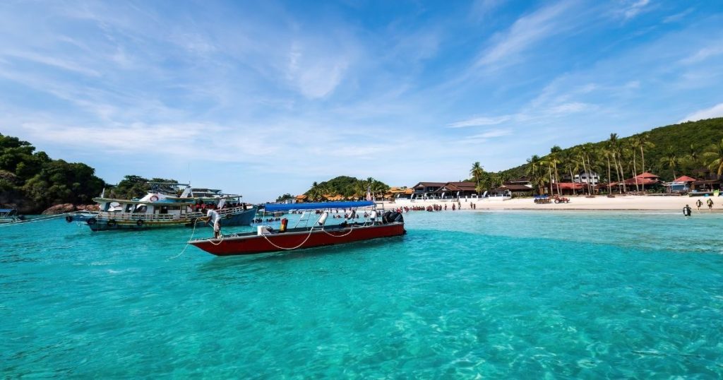 Tropical beach scene with turquoise water, a red boat near the shore, and palm trees lining a sandy beach with resort buildings in the background.