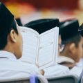 Students in traditional caps sit in chairs, reading an open book together at a ceremony or gathering.