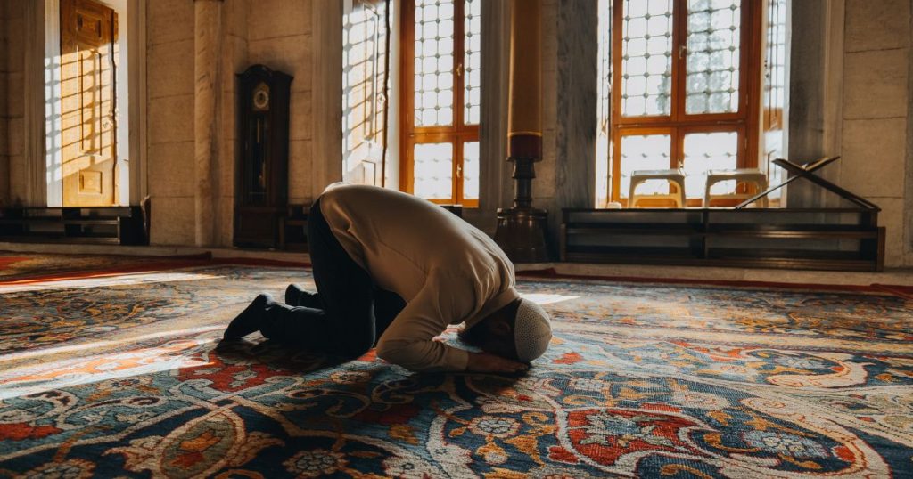 Man in a white prayer cap kneels in prostration on a richly patterned carpet inside a sunlit room, likely a mosque.